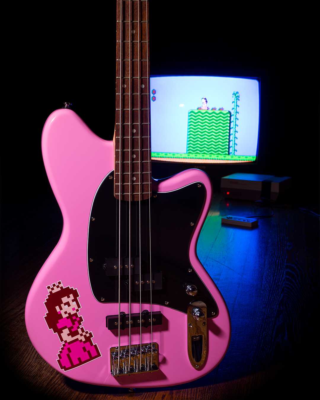 Talman Bass TMB100K-PP Peach Pink, with an 8-bit Princess Peach sticker on the front. Positined in front of an old tube TV and Nintendo Entertainment System playing Mario 2.
